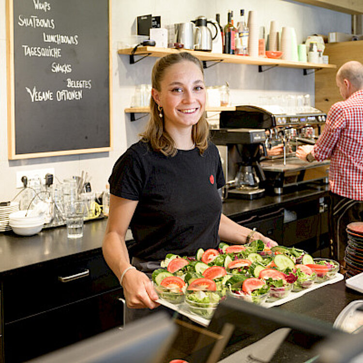 Eine junge Frau lächelt und hält ein Tablett mit frisch zubereiteten belegten Broten in einem Café. Im Hintergrund arbeitet ein Mann an der Kaffeemaschine.