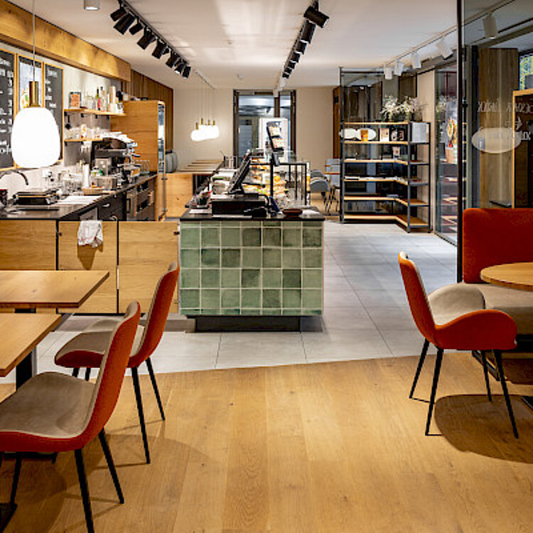 Modern eingerichtetes Café mit Holz- und grünen Fliesenakzenten, roten Stühlen und Tischen, einer Theke mit Kaffeemaschine und Regalen im Hintergrund. Eine helle, einladende Atmosphäre lädt zum Verweilen ein.