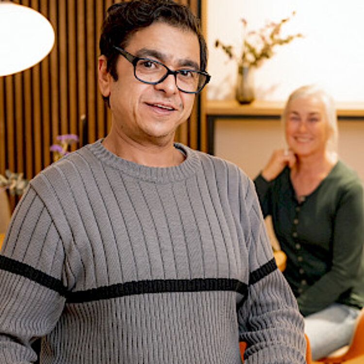 Ein Mann trägt ein Tablett mit zwei Tassen Kaffee und Keksen, im Hintergrund sitzt eine lächelnde Frau. Beide wirken entspannt und freundlich.