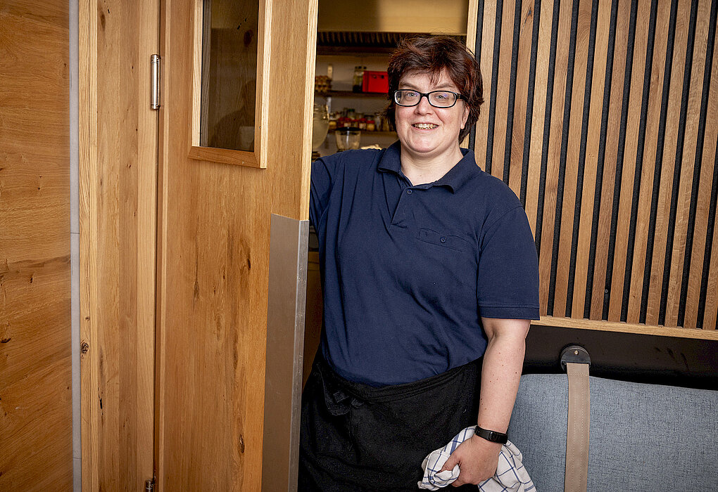 Frau in Arbeitskleidung öffnet lächelnd eine Holztür und hält ein Geschirrtuch in der Hand, in einem Raum mit Holzwänden und modernem Interieur.