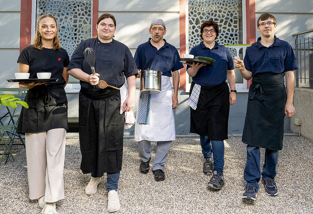 Fünf lächelnde Servicekräfte in Arbeitskleidung gehen gemeinsam auf die Kamera zu, dabei tragen sie verschiedene Küchenutensilien und Speisen.