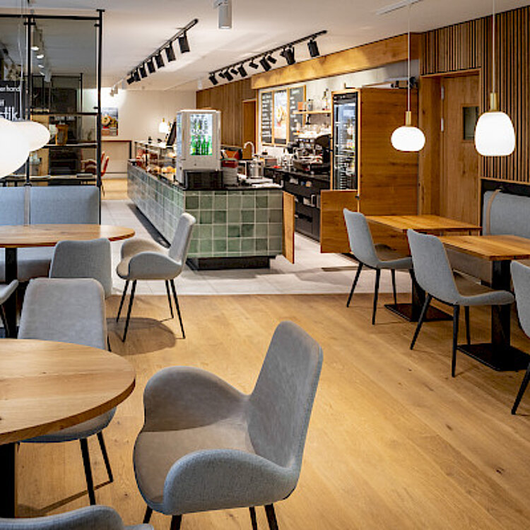 Heller, moderner Café-Innenraum mit Holzboden, diversen grauen Stühlen und Holztischen, gepolsterten Sitzbänken an der Wand und einer langen Theke im Hintergrund. Deckenlampen schaffen eine gemütliche Atmosphäre.