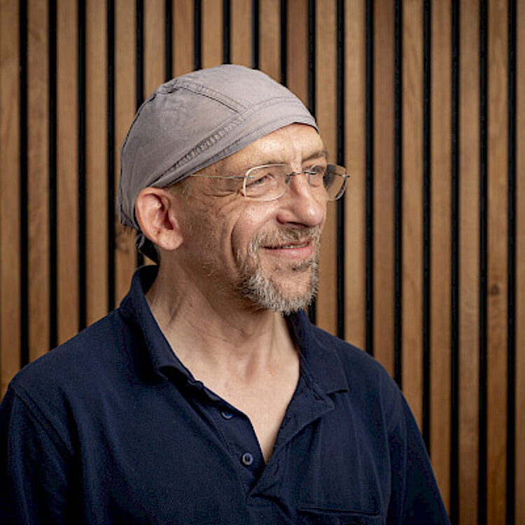 Porträt eines lächelnden älteren Mannes mit Brille und grauer Kopfbedeckung vor Holzlatten-Wand.