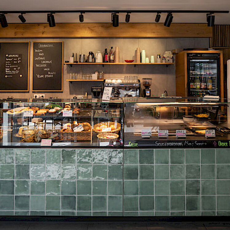 Theke einer Bäckerei oder eines Cafés mit verschiedenen Backwaren und Getränken. Im Hintergrund hängen Tafeln mit Getränkekarten und Preisen.