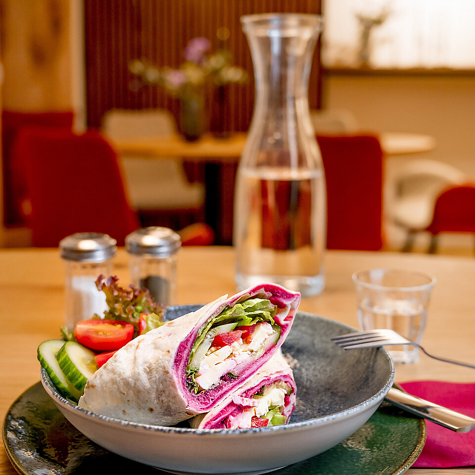 Ein Teller mit zwei aufgeschnittenen Wraps, gefüllt mit frischem Gemüse und Käse, serviert mit Gurkenscheiben, Tomaten und Salat. Im Hintergrund ist eine Karaffe mit Wasser und ein Glas zu sehen, situiert in einem gemütlichen Café.