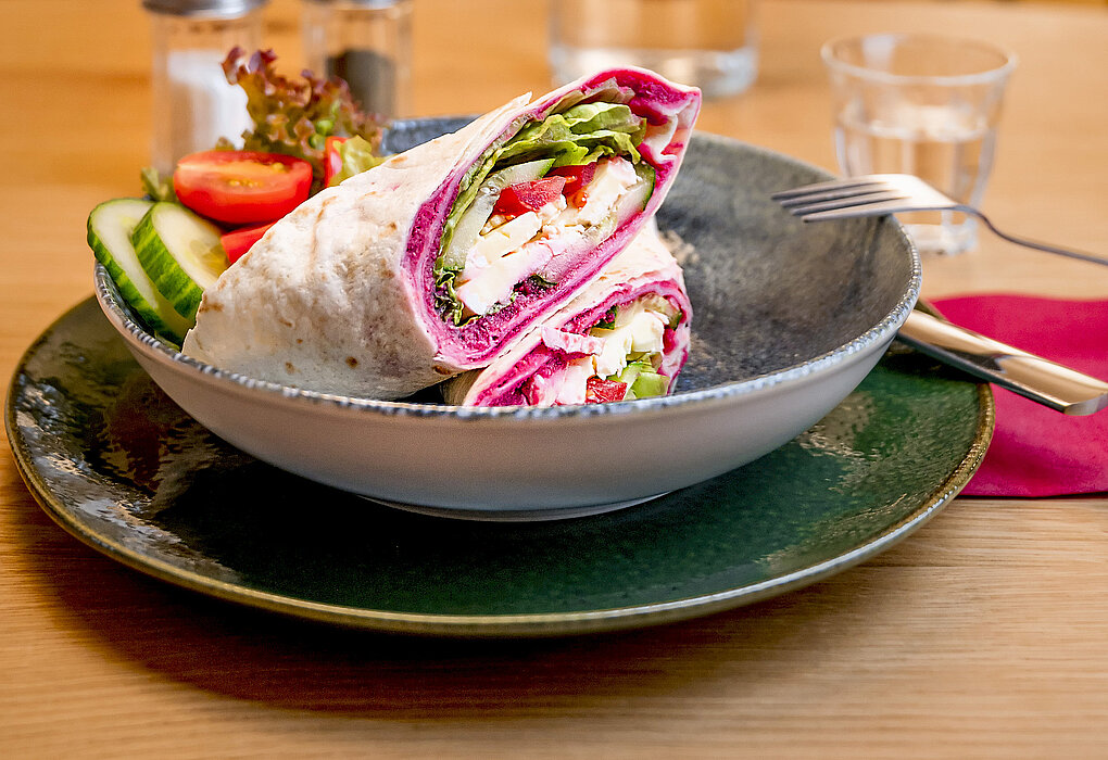 Ein Teller mit zwei aufgeschnittenen Wraps, gefüllt mit frischem Gemüse und Käse, serviert mit Gurkenscheiben, Tomaten und Salat. Im Hintergrund ist eine Karaffe mit Wasser und ein Glas zu sehen, situiert in einem gemütlichen Café.