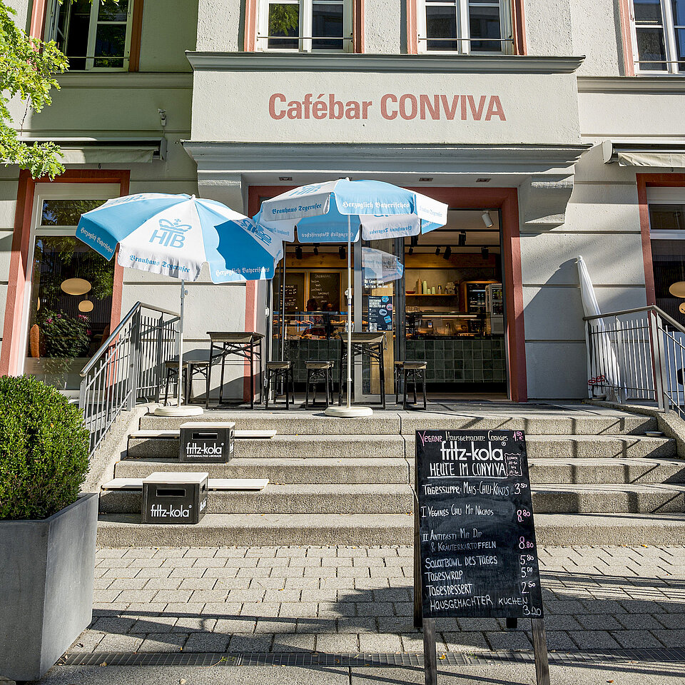 Außenansicht einer Cafebar namens „CONVIVA“ mit Tischen, Stühlen und blauen Sonnenschirmen auf der Treppe, sowie einer Tafel mit Tagesangeboten vor dem Eingang.