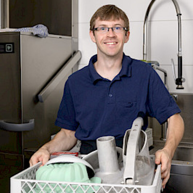 Ein lächelnder Mann in Arbeitskleidung hält eine Spülkiste mit sauberem Geschirr in einer industriellen Küchenumgebung.