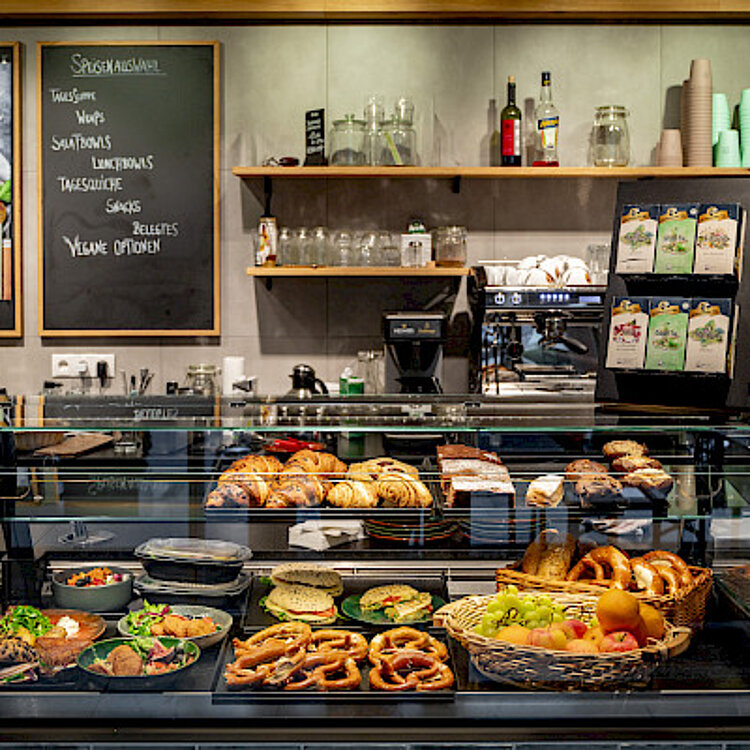 Eine Vitrine in einem Café oder Bistro, gefüllt mit verschiedenen frischen Backwaren wie Brezeln, Croissants und belegten Brötchen, sowie Obst. Im Hintergrund sind Regale mit Gläsern, Kaffee- und Teezubehör zu sehen. Eine Tafel und ein Menü weisen auf Speisen hin.